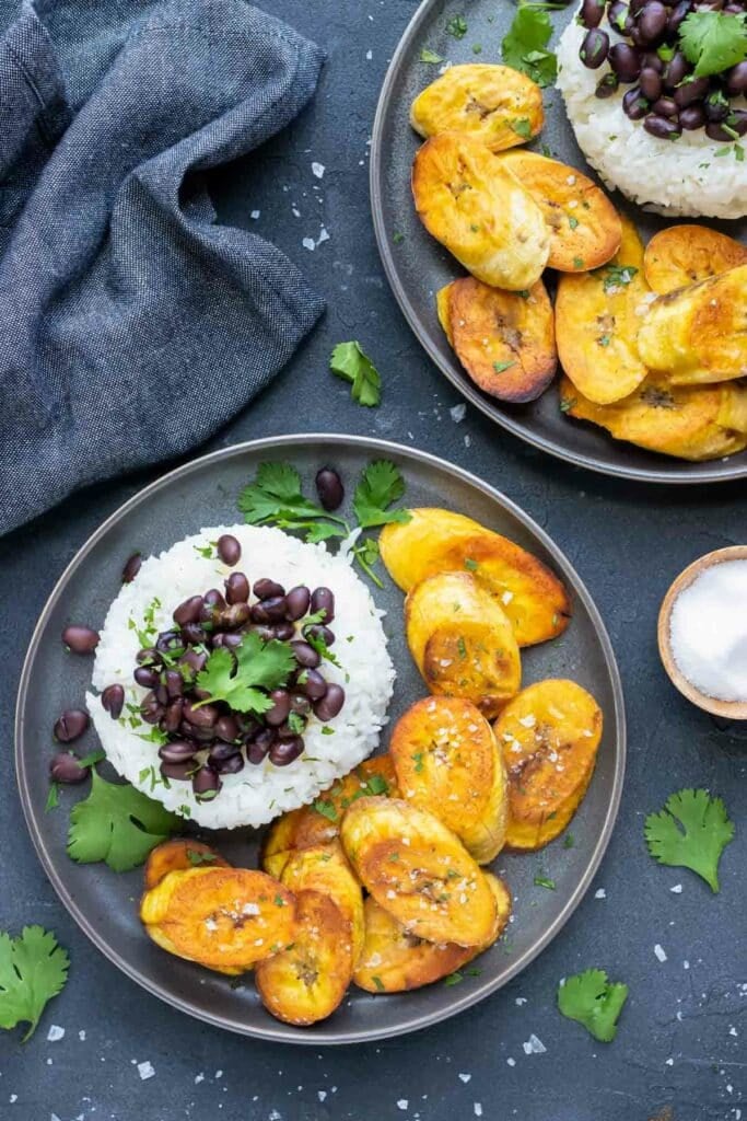 Sweet, ripe, fried plantains on a grey plate next to white rice and black beans.