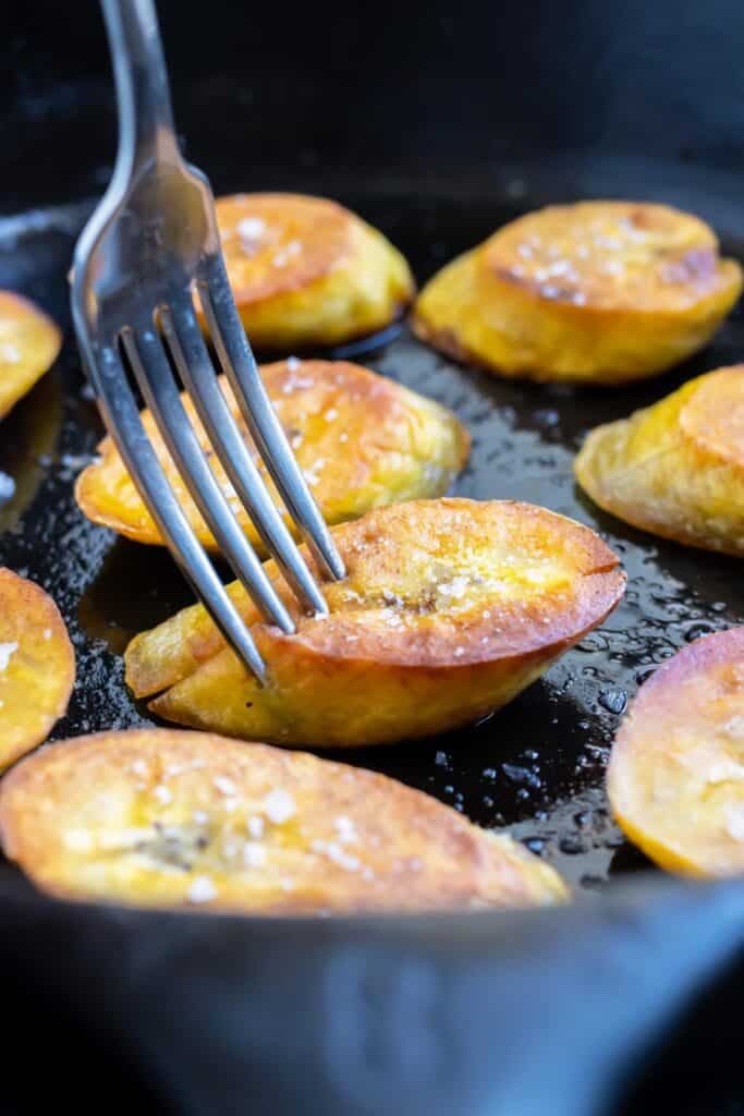 A fork picking up a plantains that has been fried in a skillet.
