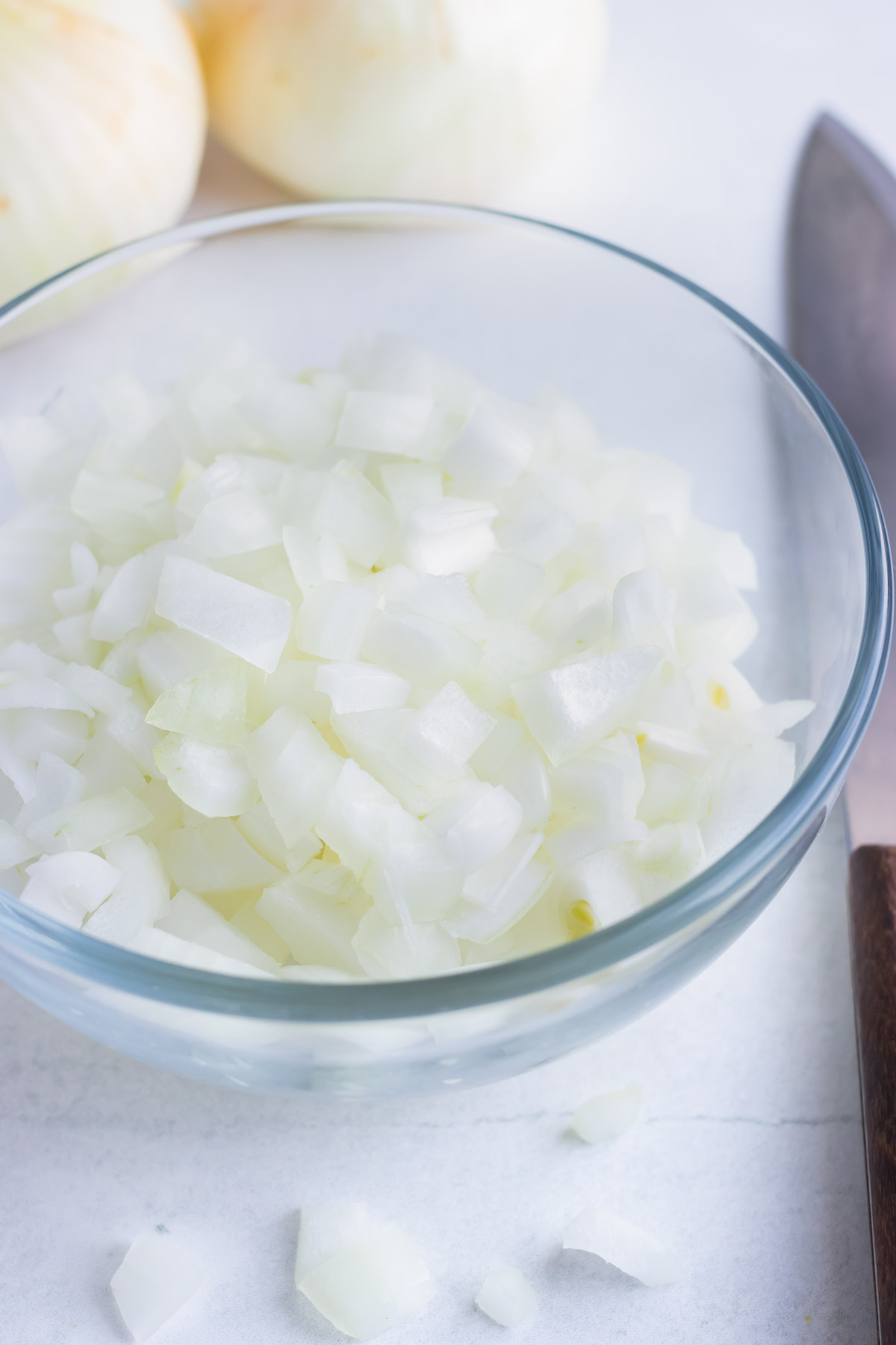 A clear bowl of chopped onions sits on a white cutting board next to a sharp knife.