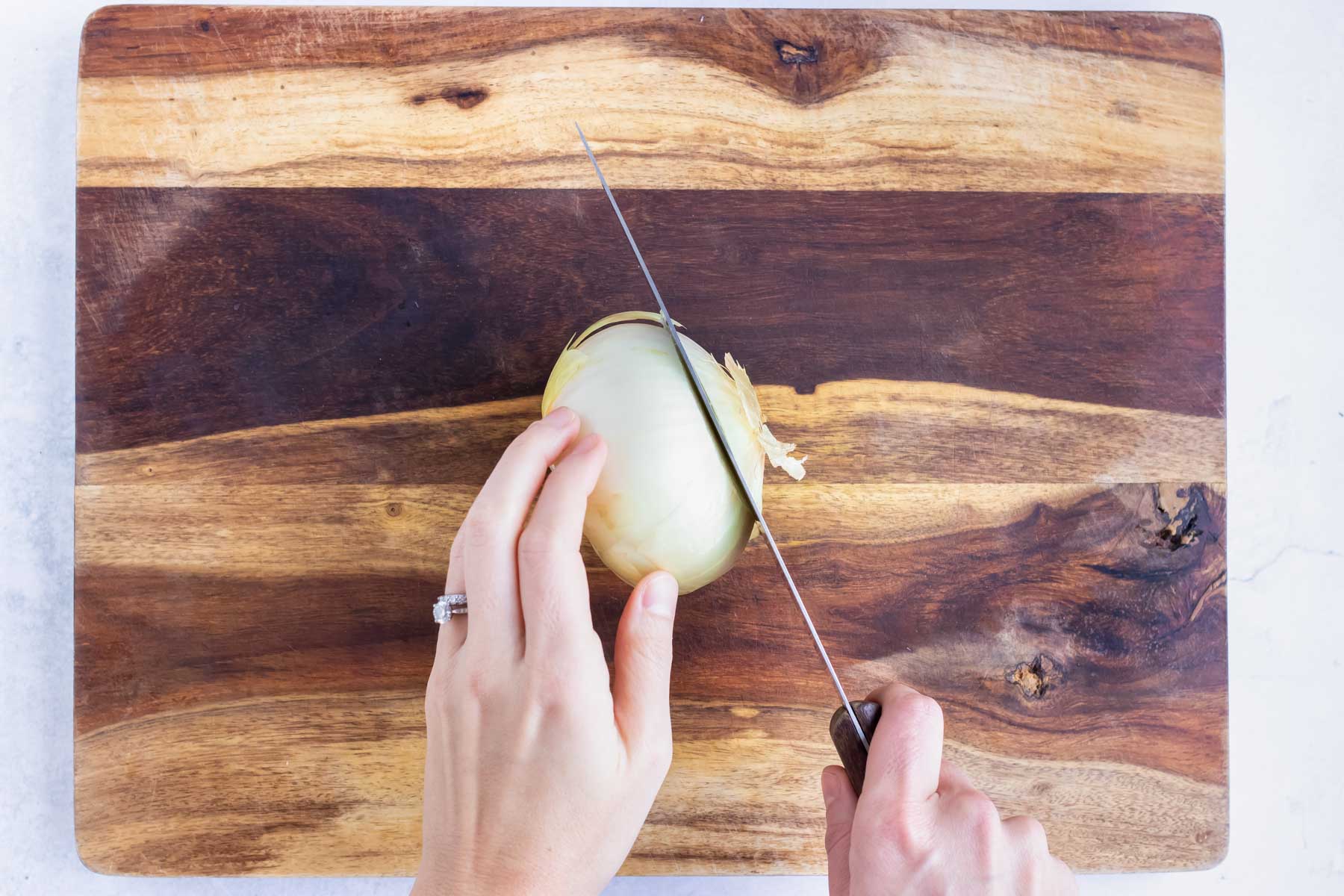 The end of an onion is removed with a knife on a cutting board.