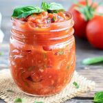 A glass jar full of a spaghetti sauce recipe with tomatoes and garlic in the background.