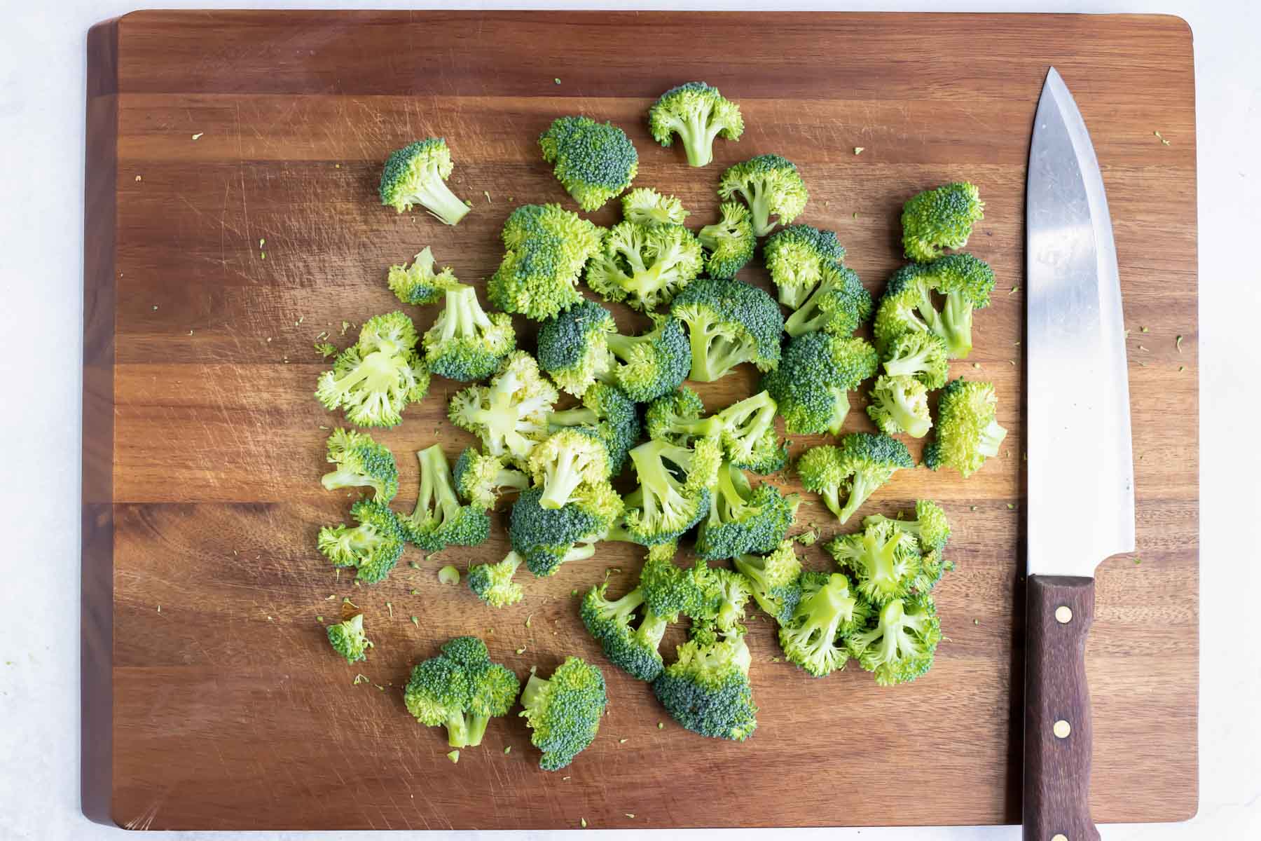 Broccoli florets are chopped on a wooden cutting board.