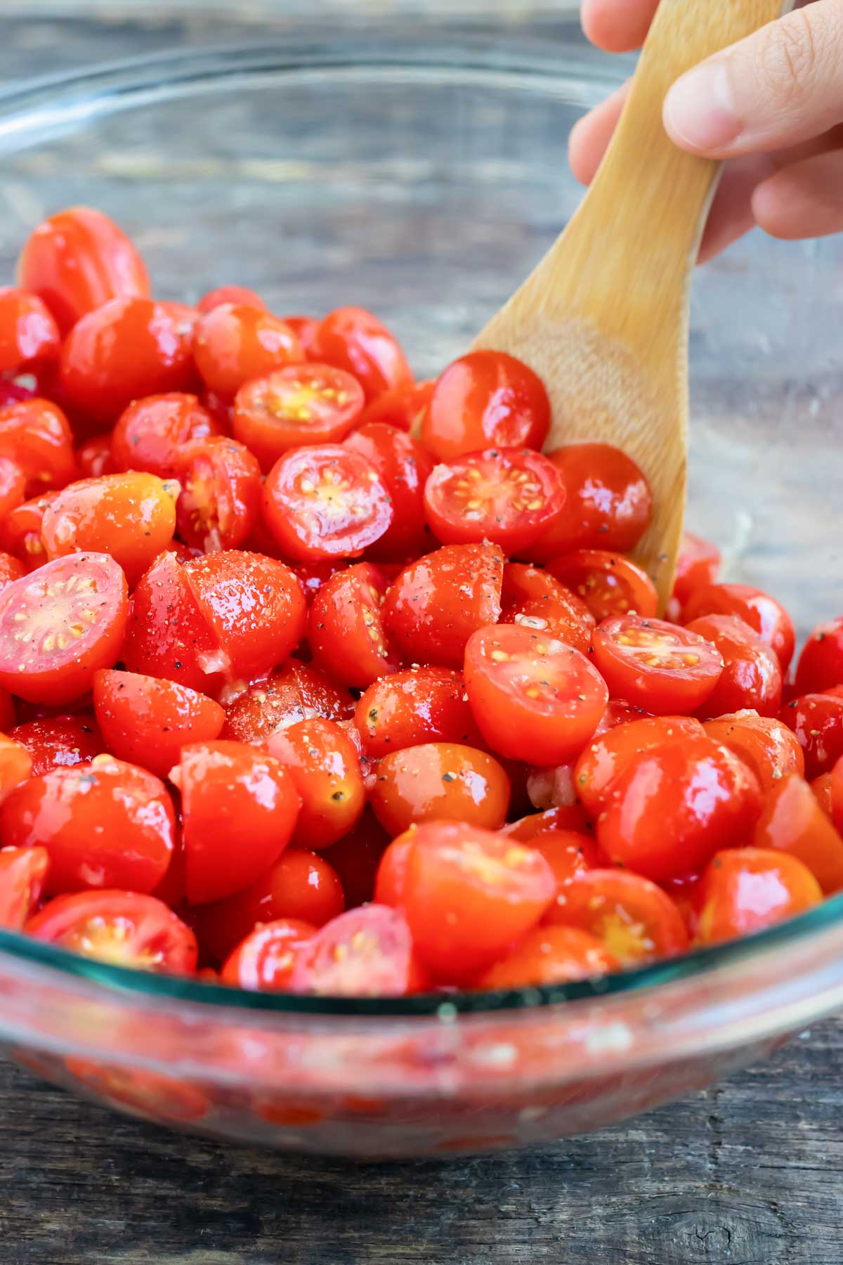 A wooden spoon stirs sliced tomatoes with seasoning.