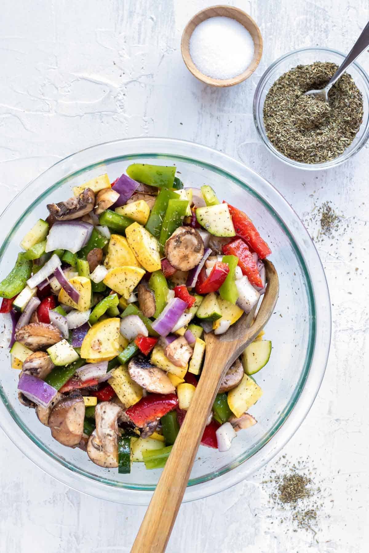 A glass bowl full of chopped zucchini, squash, bell peppers, and onions next to a bowl full of Italian seasoning and salt.