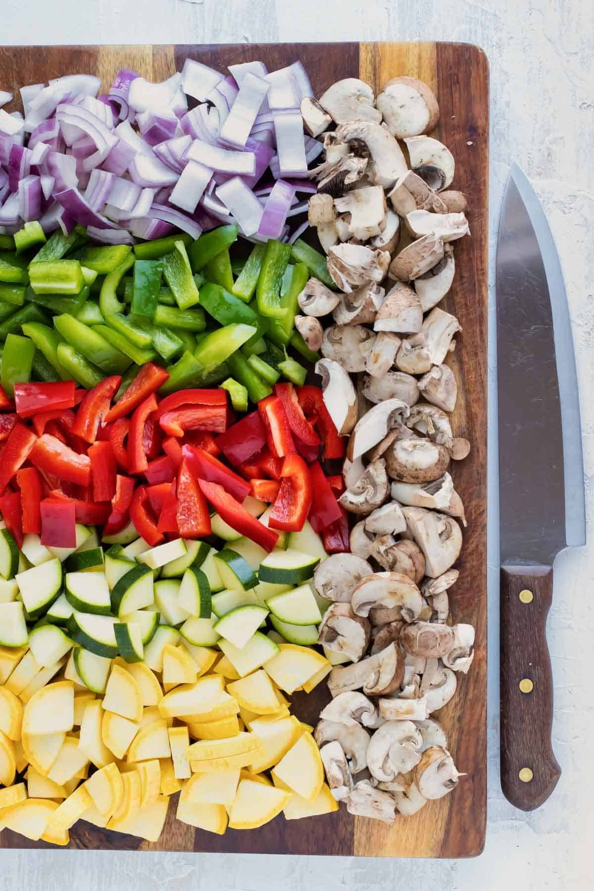 A cutting board full of yellow squash, zucchini squash, red onion, green and red bell pepper, and mushrooms.