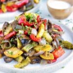 A pile of oven-roasted vegetables on a white plate with another serving in the background.
