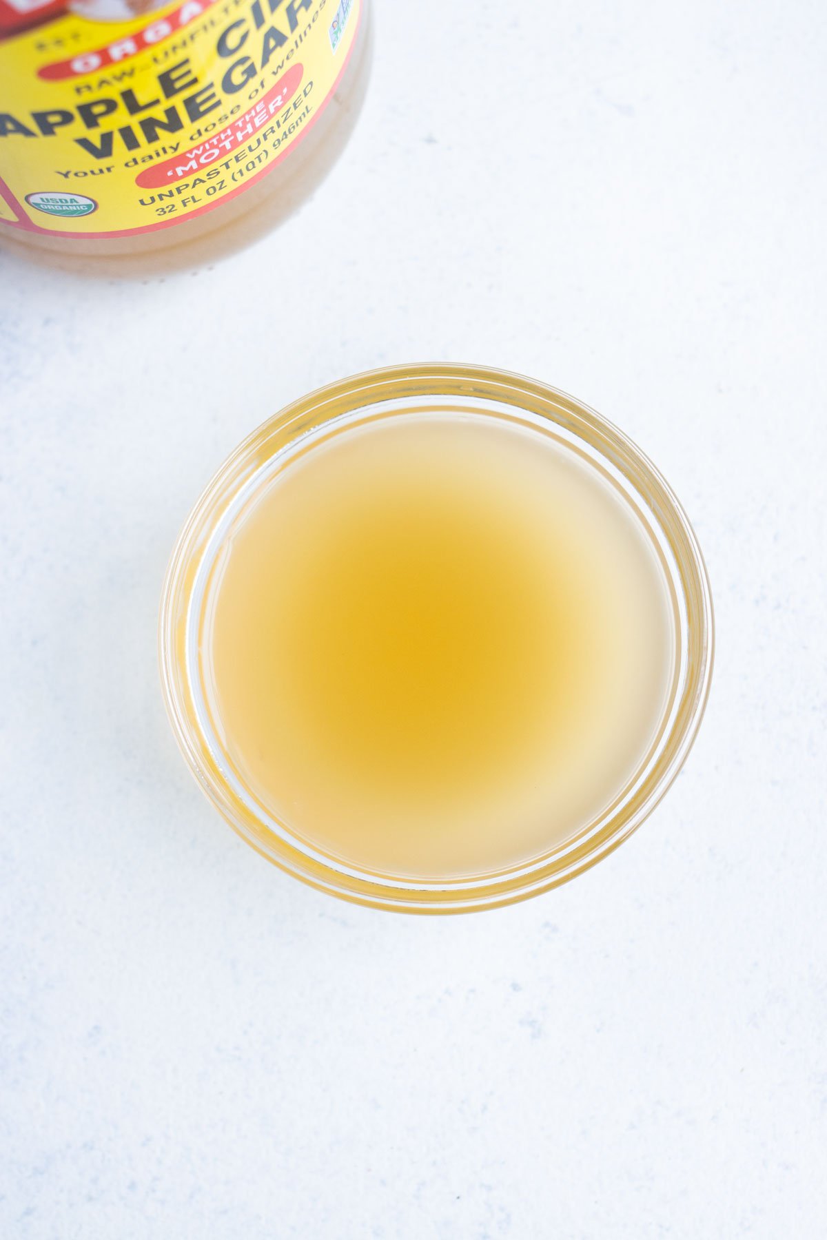 Bird's eye view of apple cider vinegar in a small glass bowl with the container in the background.