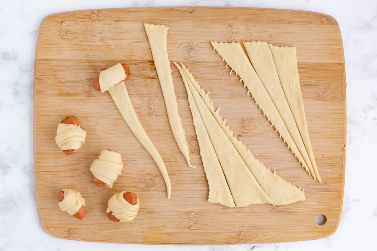 A cutting board with sliced crescent rolls and rolled pigs in a blanket.