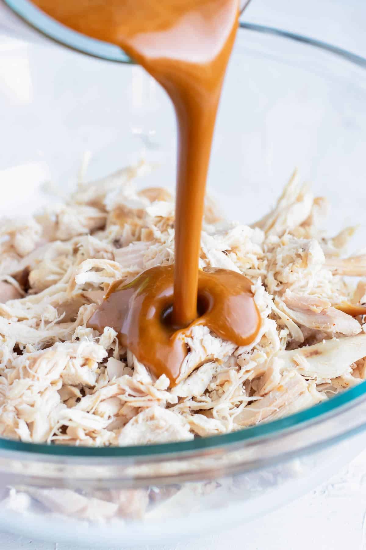 Asian peanut butter sauce being poured into a clear bowl full of shredded rotisserie chicken.