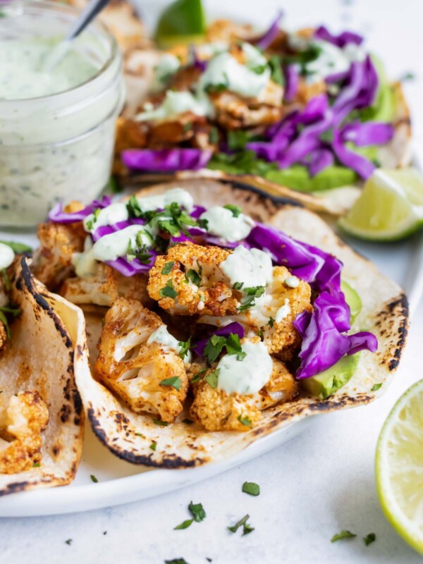 A dinner tray serving roasted cauliflower tacos with a jar full of vegan cilantro cream sauce in the middle with limes.