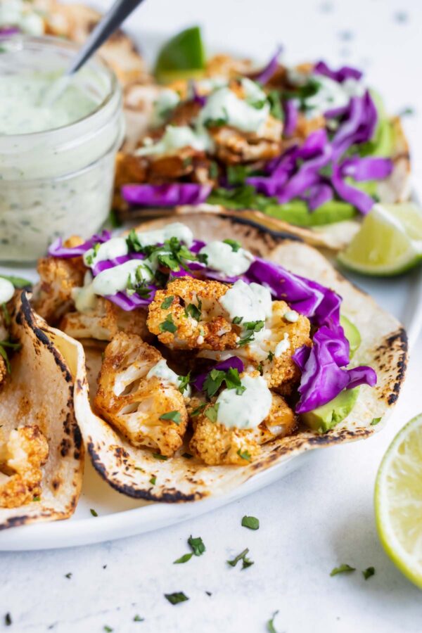 A dinner tray serving roasted cauliflower tacos with a jar full of vegan cilantro cream sauce in the middle with limes.