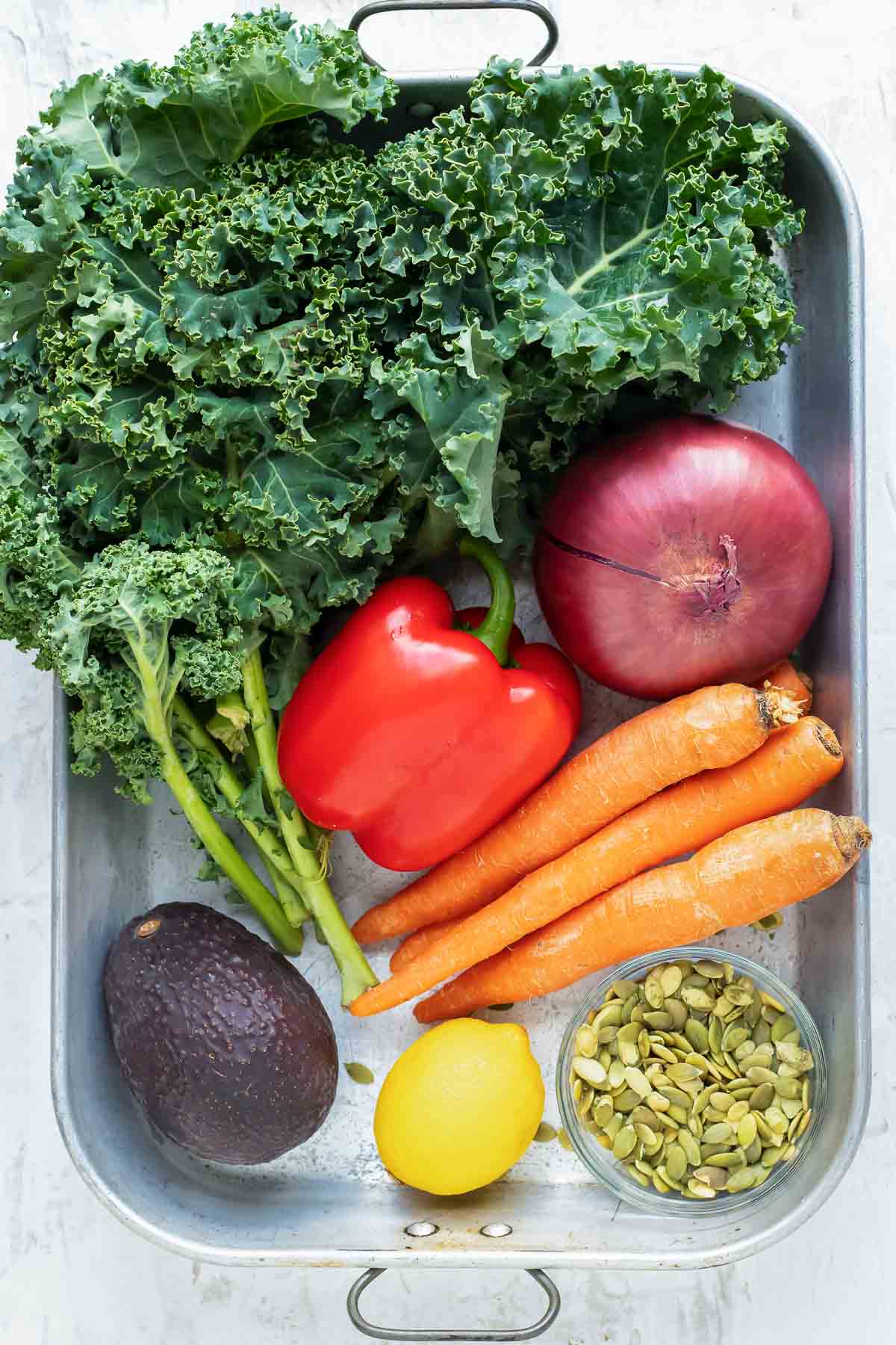 A bunch of kale, red bell pepper, avocado, lemon, pumpkin seeds, and carrots in a silver basket as the ingredients for a massaged kale salad.