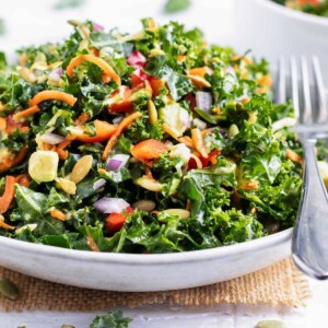 A healthy kale salad recipe with carrots, avocado, and a lemon salad dressing in a white bowl.