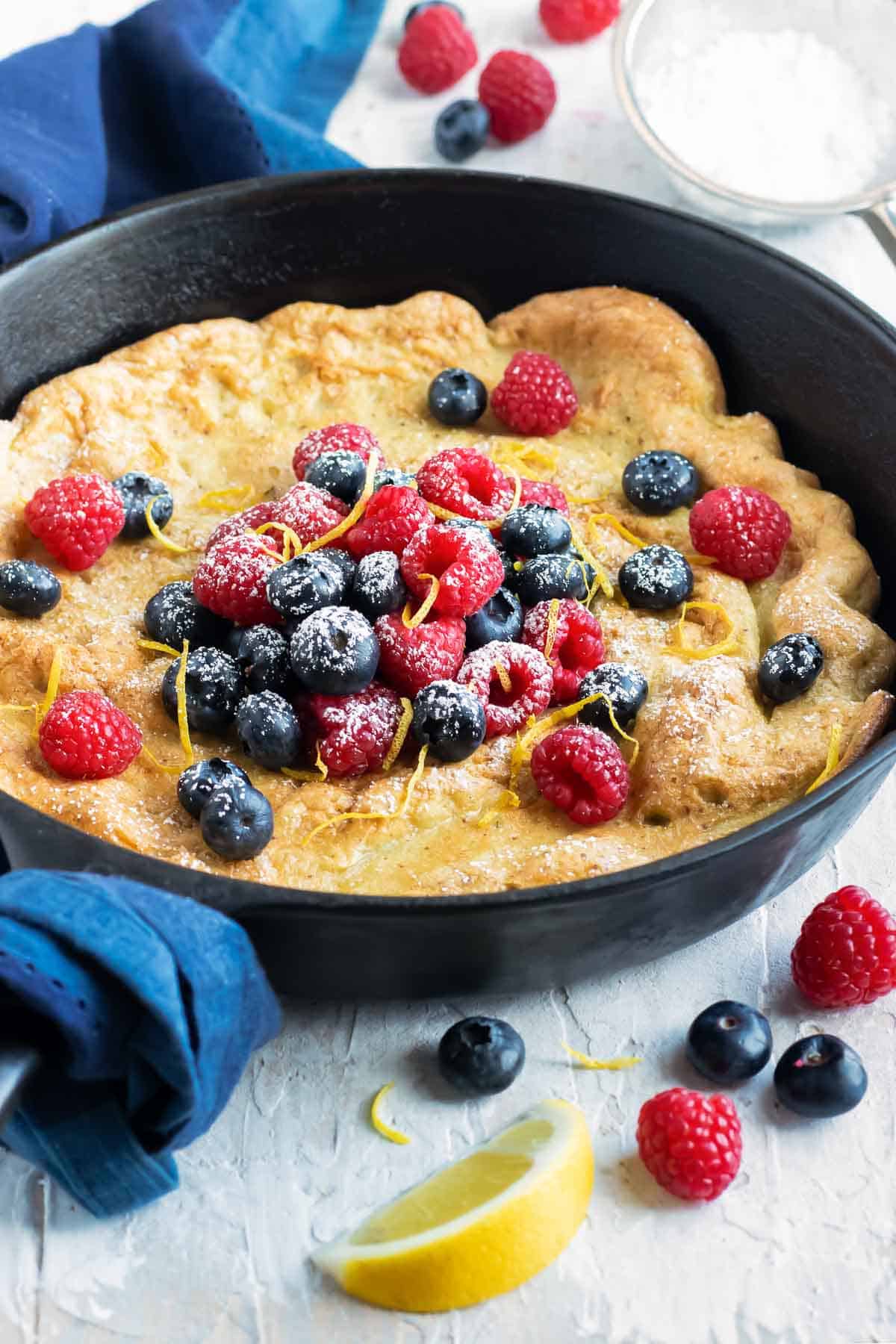 A skillet full of a Dutch baby popover pancake recipe surrounded by fresh berries and lemons.