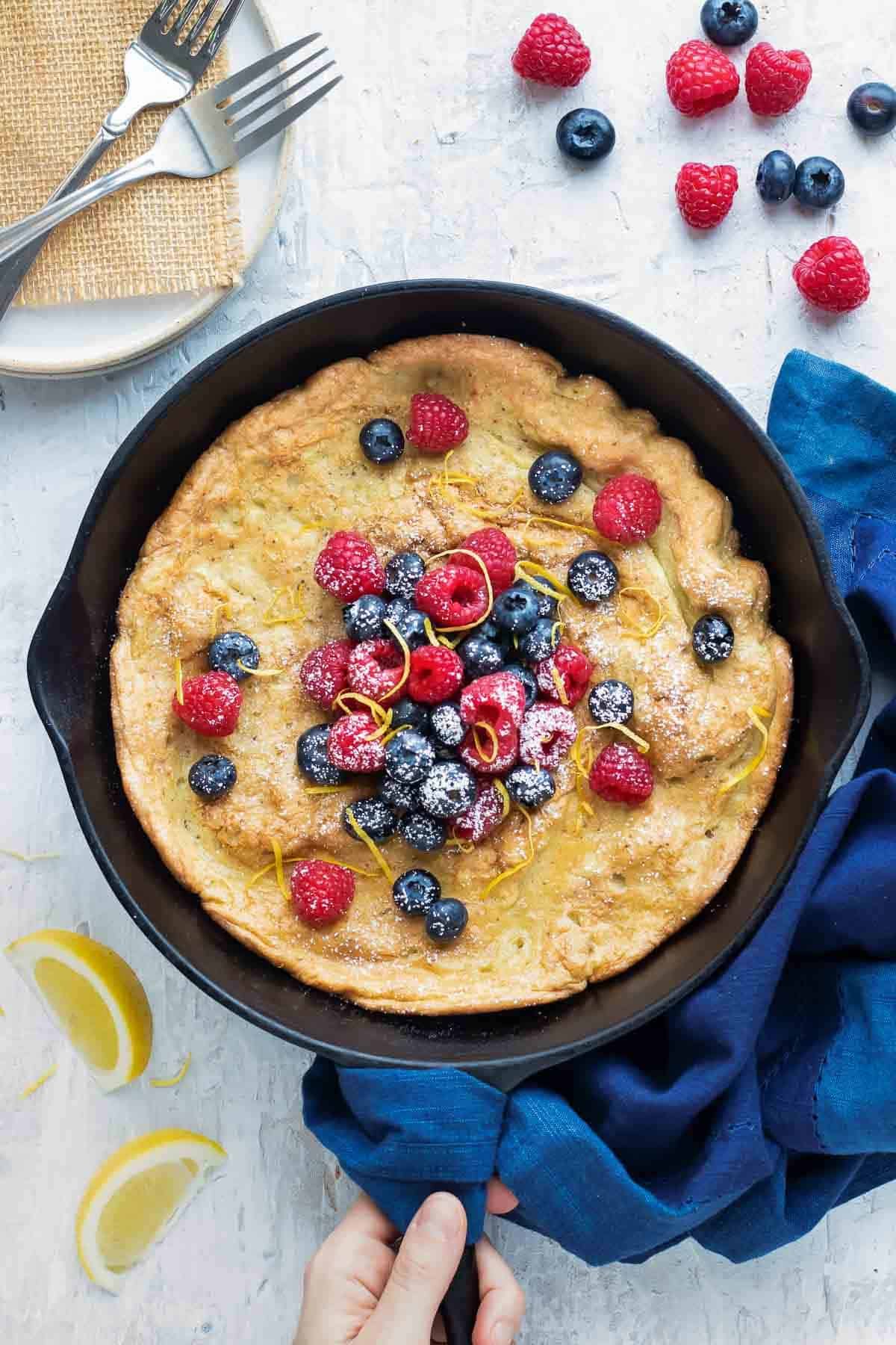 A hand holding a cast iron skillet with a Dutch baby, or German pancake, with two plates and two forks.