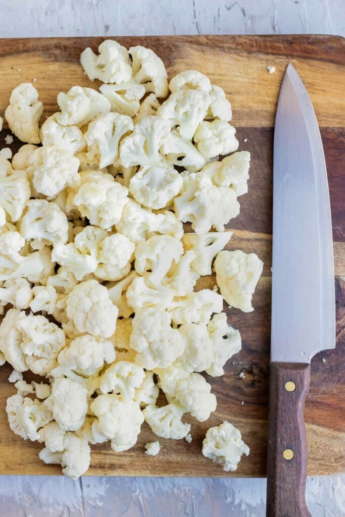 Small cauliflower florets on a wooden cutting board next to a chef's knife.