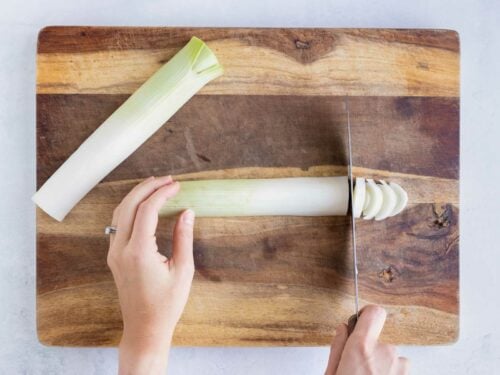 A cutting board with a fresh leek on it and cutting it into thin slices.