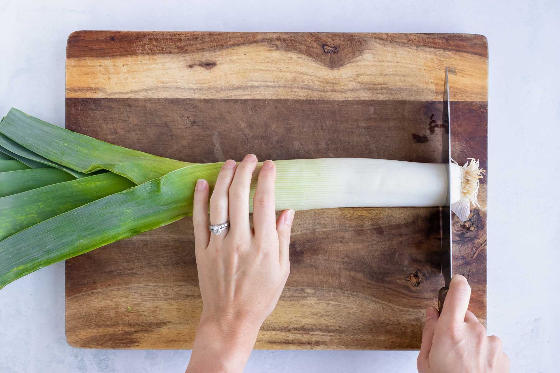 One leek on a wooden cutting board with its root being cut off with a sharp knife.