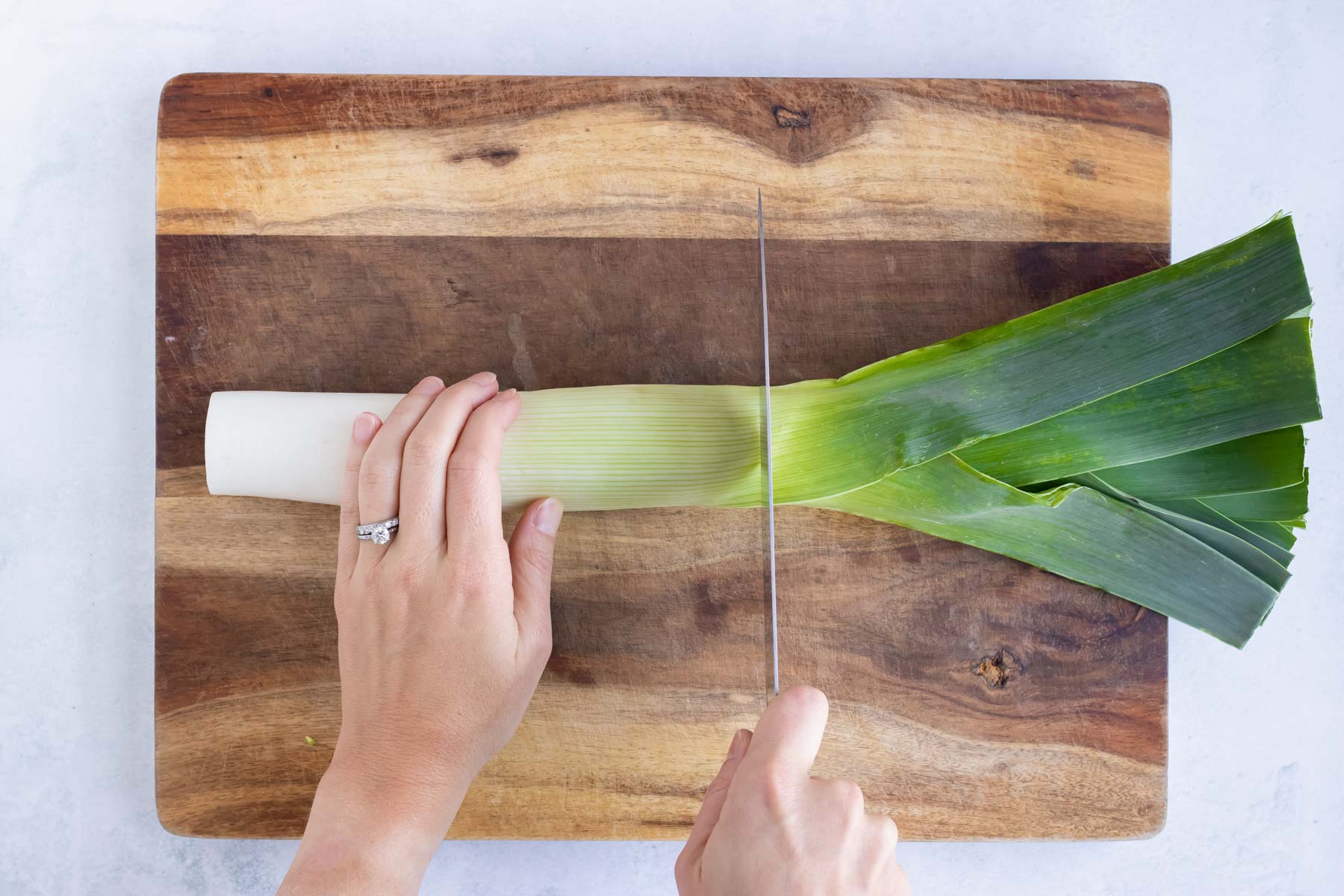 The green leaves of a leek being cut off and discarded on a wooden cutting board.