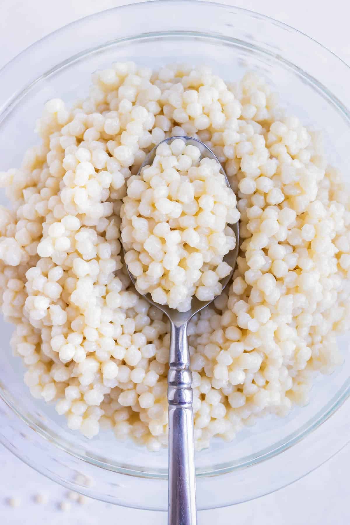 A spoon scooping up some cooked pearl couscous from a glass bowl.