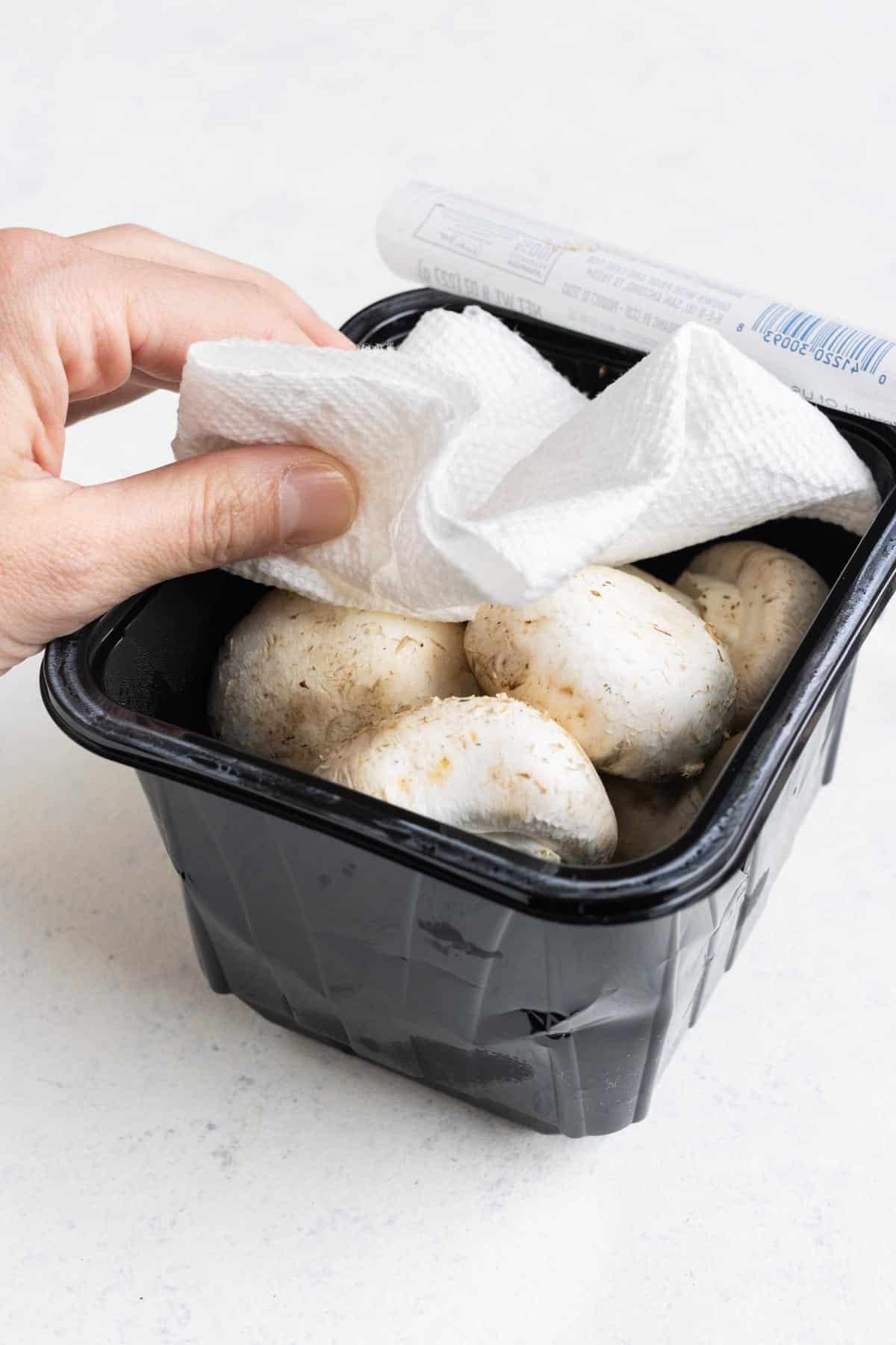 Putting a paper towel on top of a store-bought container full of whole mushrooms.