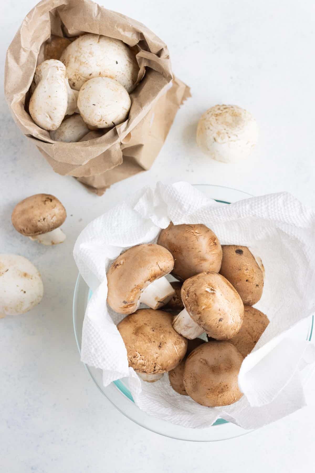 One brown paper bag full of whole mushrooms and a colander bowl of whole mushrooms with a paper towel.