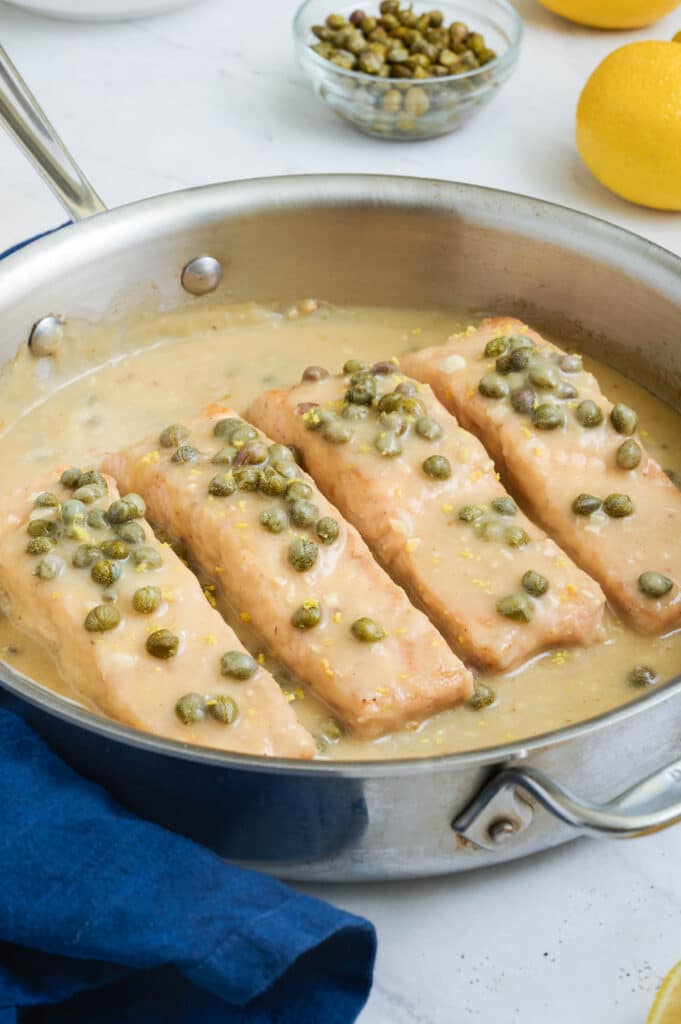 Salmon piccata filets are in a skillet with capers and a lemon garlic butter sauce.