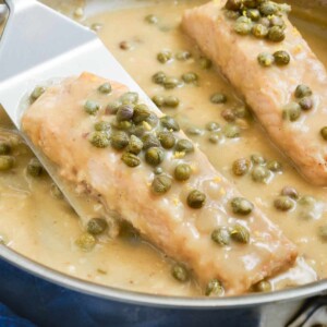 A spatula picks up a filet of salmon piccata with a creamy lemon butter sauce from a pan.