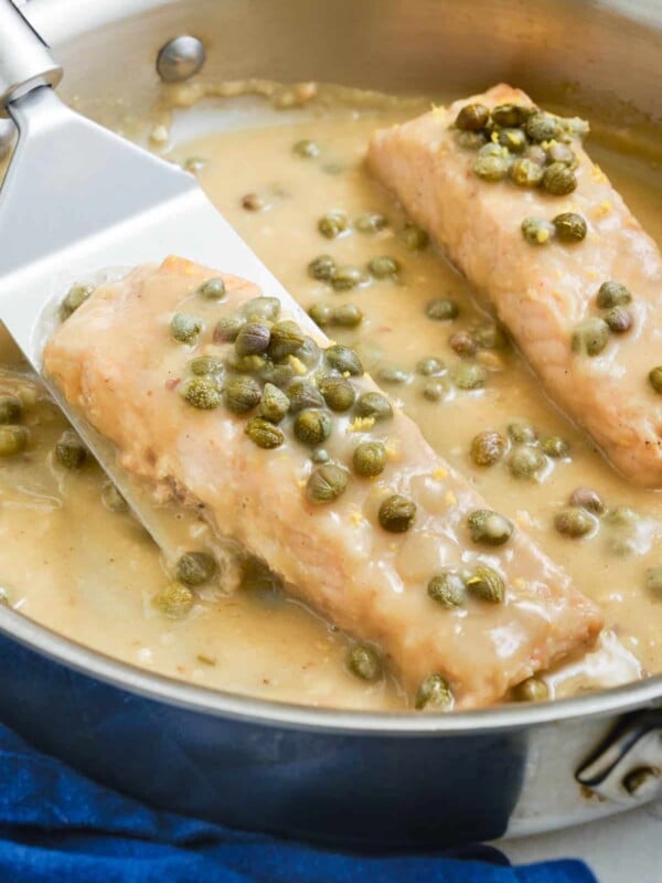 A spatula picks up a filet of salmon piccata with a creamy lemon butter sauce from a pan.