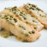 Salmon filets in piccata sauce are served on a plate with capers.