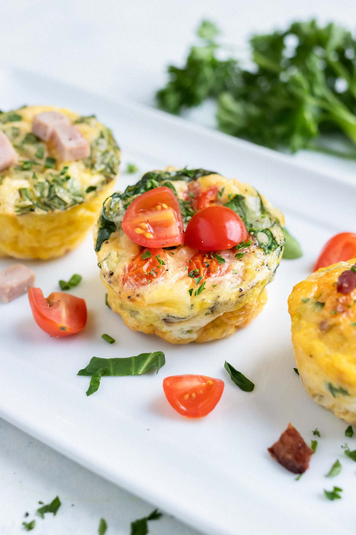 Three breakfast egg muffins are served one a plate with fresh tomatoes and herbs.