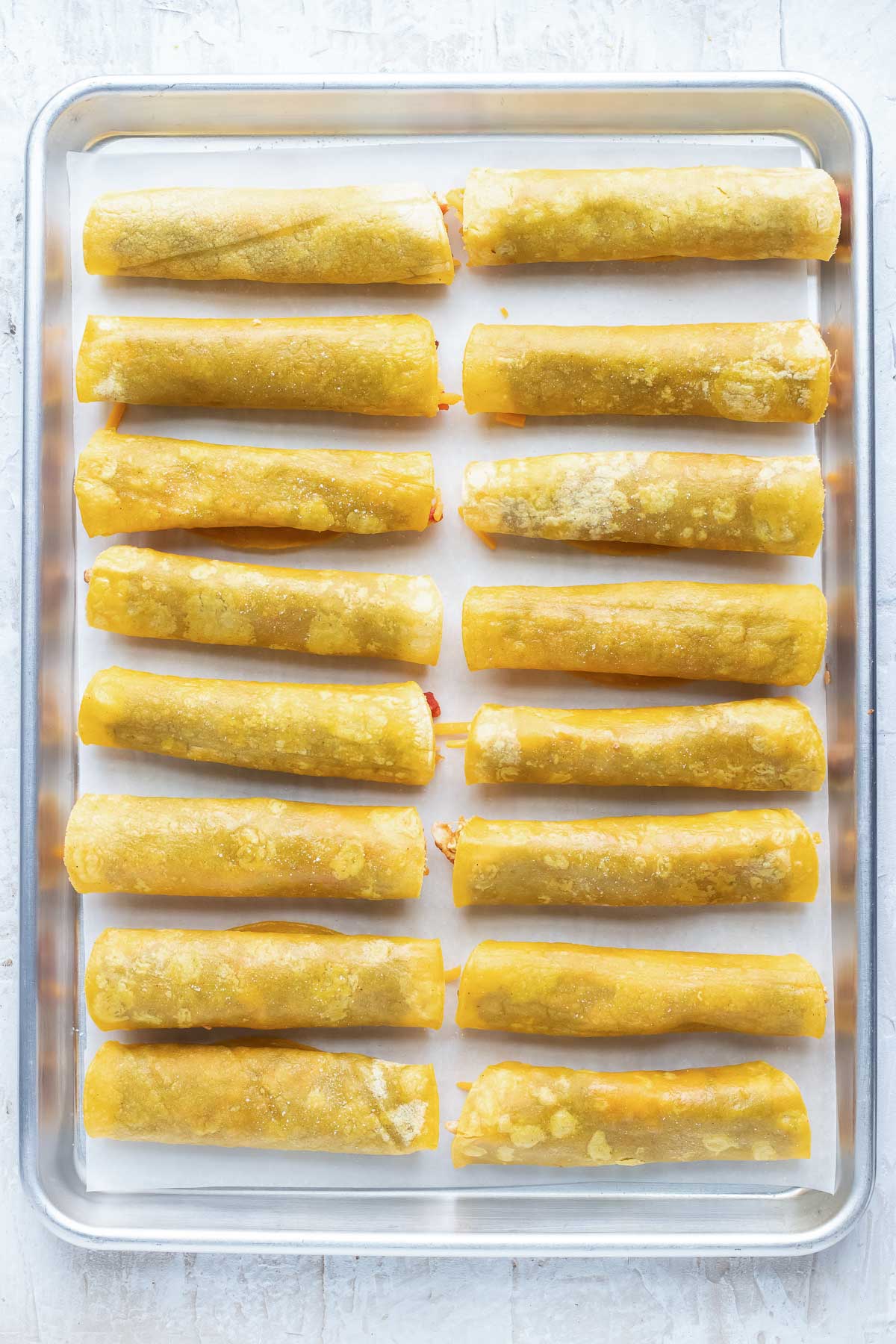 Sixteen taquitos rolled up and placed seam side down on a parchment paper lined baking sheet.