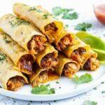 A stack of baked chicken taquitos with refried beans and cheese on a white plate for a Cinco de Mayo party.