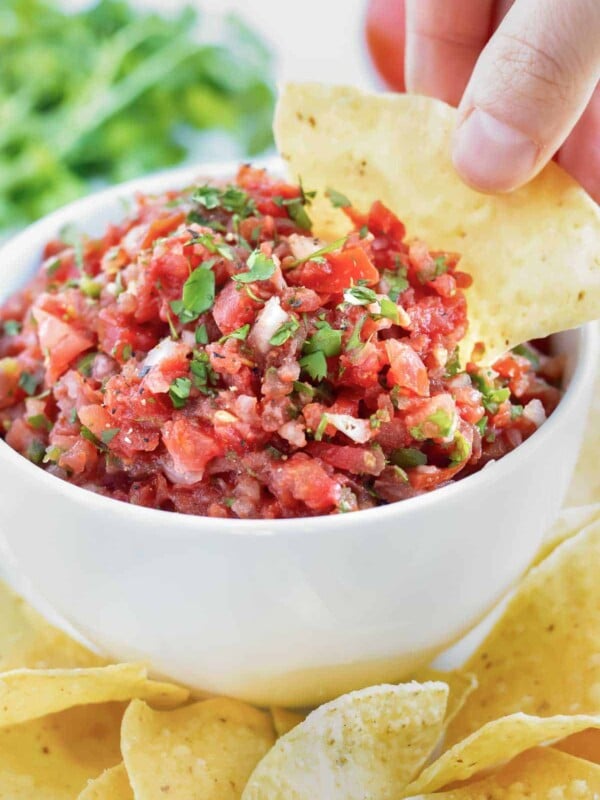 A thick and chunky salsa in a white bowl and a hand dipping a tortilla chip into it.