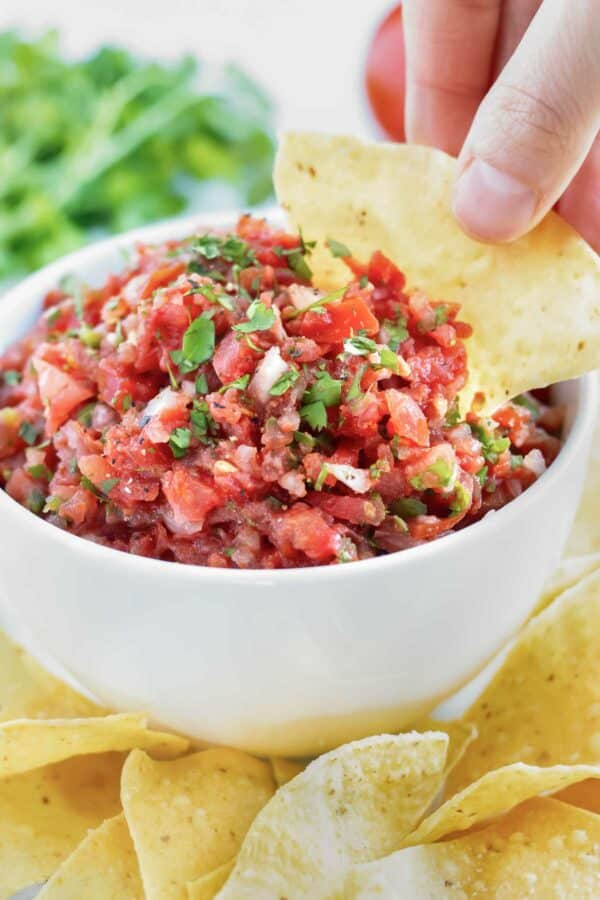 A thick and chunky salsa in a white bowl and a hand dipping a tortilla chip into it.