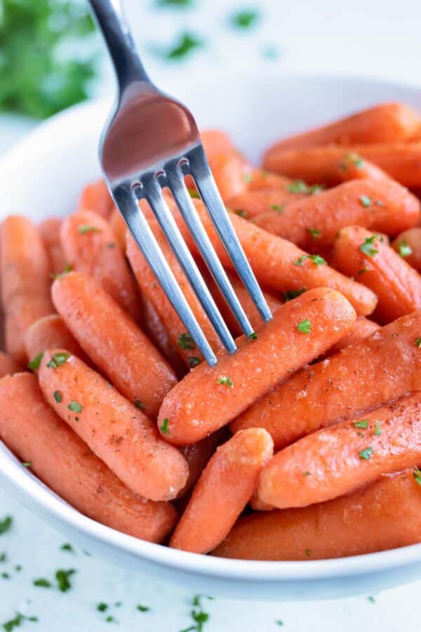 A baby carrot is eaten with a metal fork from a bowl of glazed carrots.