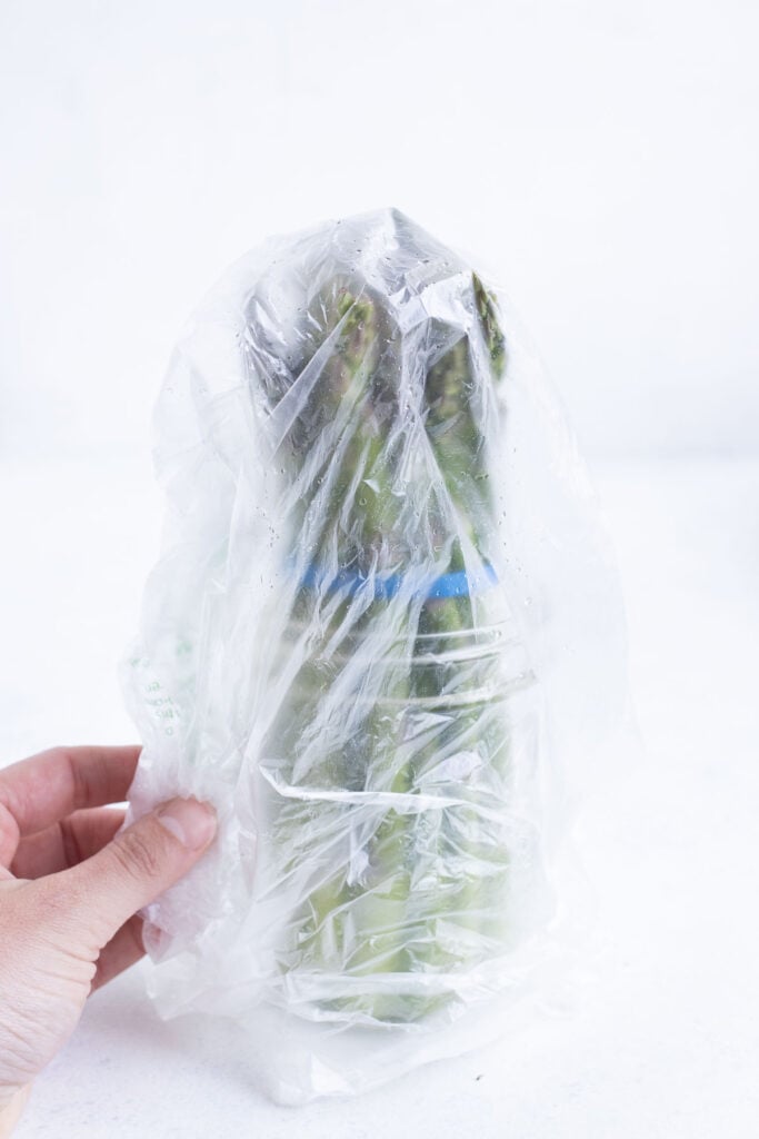 One bunch of asparagus sitting in a mason jar with water and plastic wrap loosely placed over the top of the asparagus spearheads.