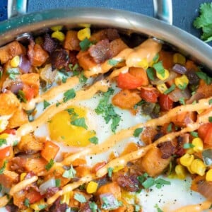 A stainless steel skillet full of sweet potato hash browns with eggs and sriracha mayo.