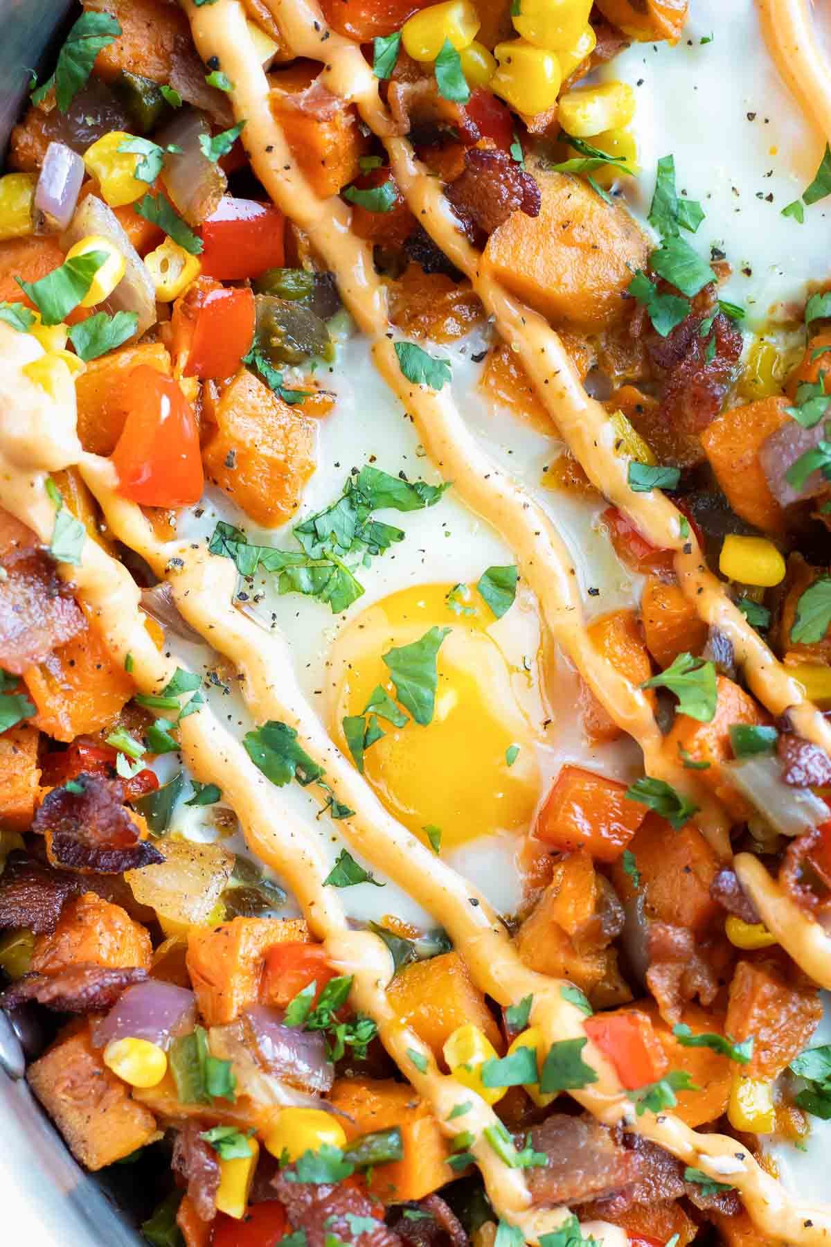 An egg in the middle of a sweet potato hash with sriracha mayo and cilantro on top.