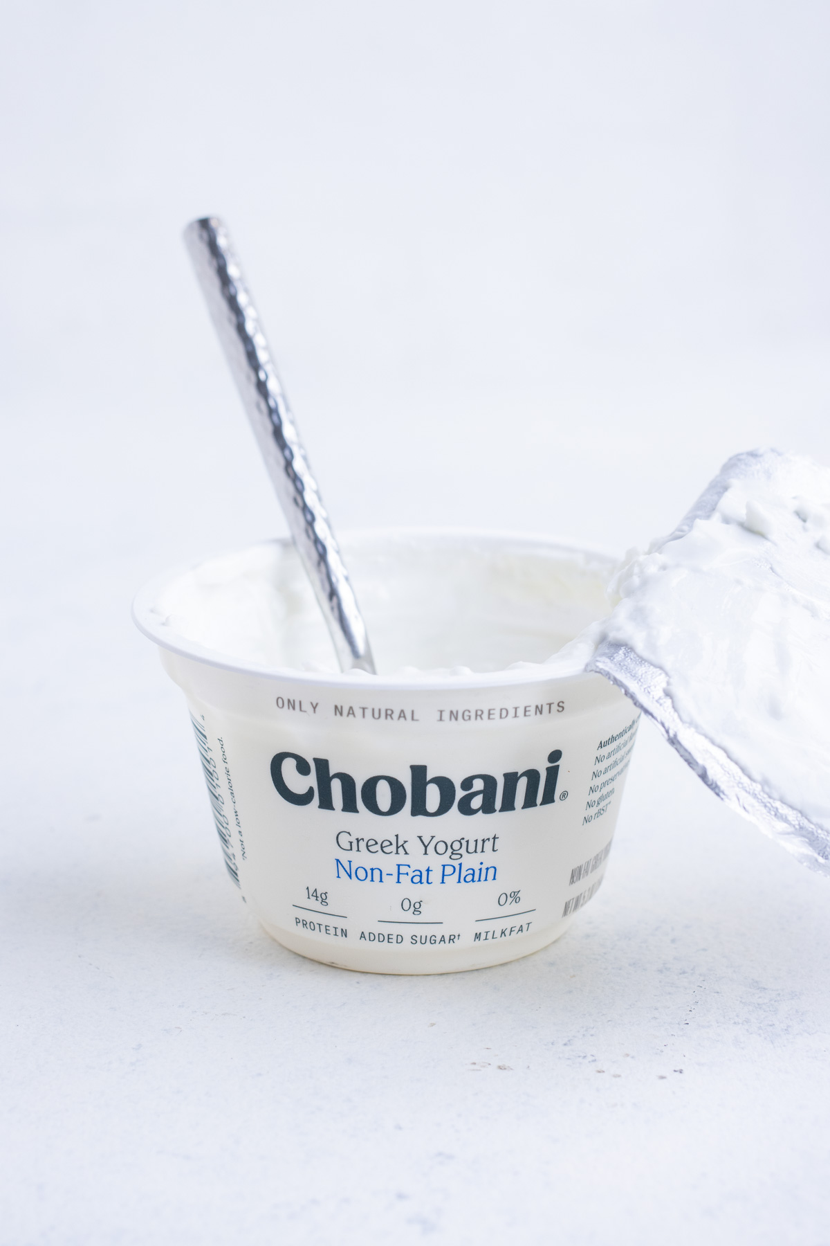 An opened plain Greek yogurt container with its lid ripped off with a spoon inside.