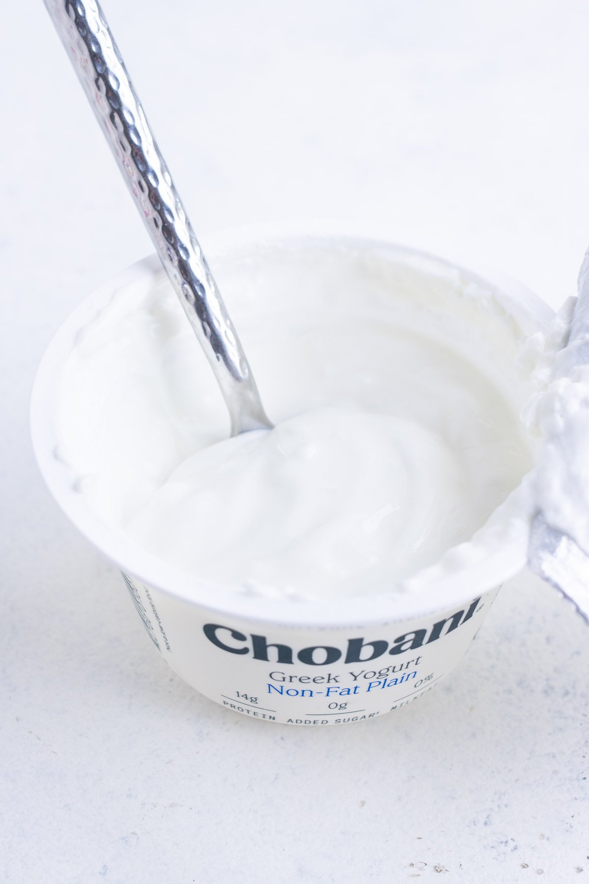 An opened plain Greek yogurt container with its lid ripped off with a spoon inside.