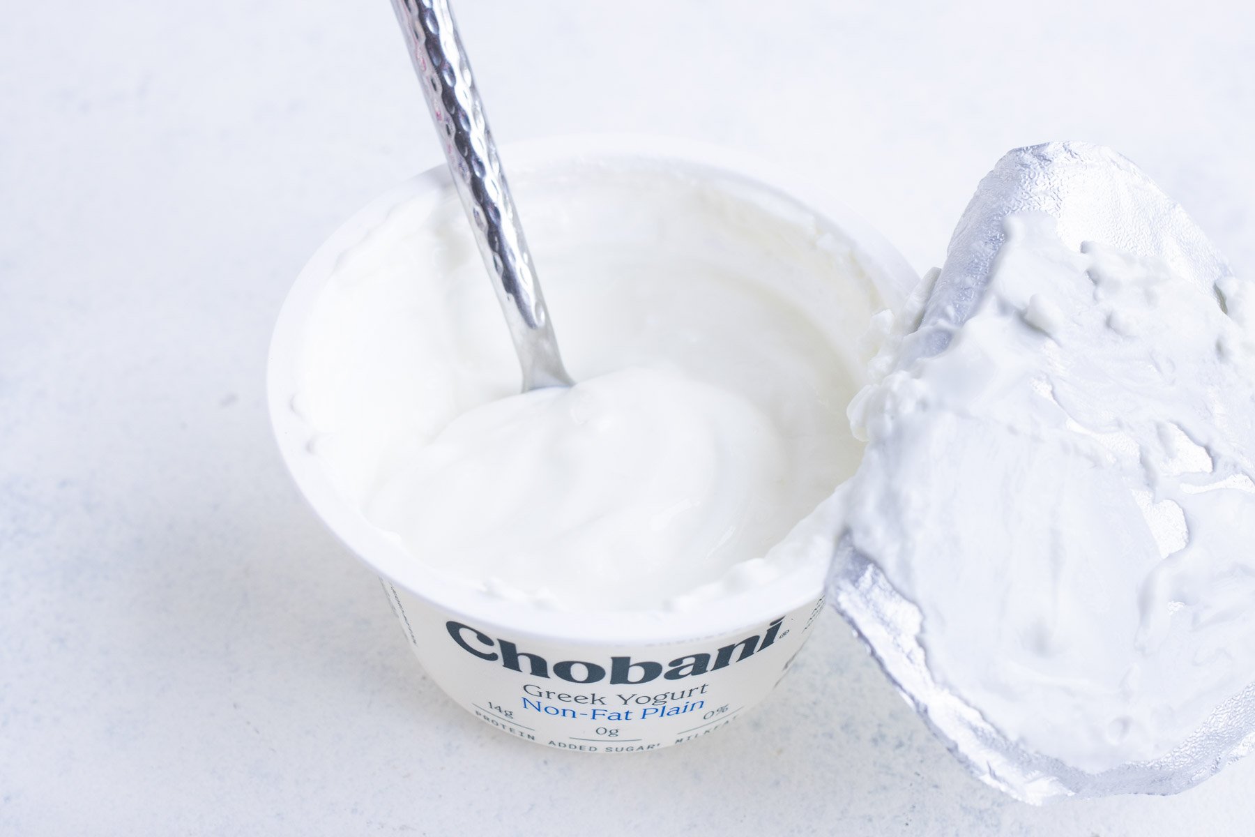 An opened plain Greek yogurt container with its lid ripped off with a spoon inside.