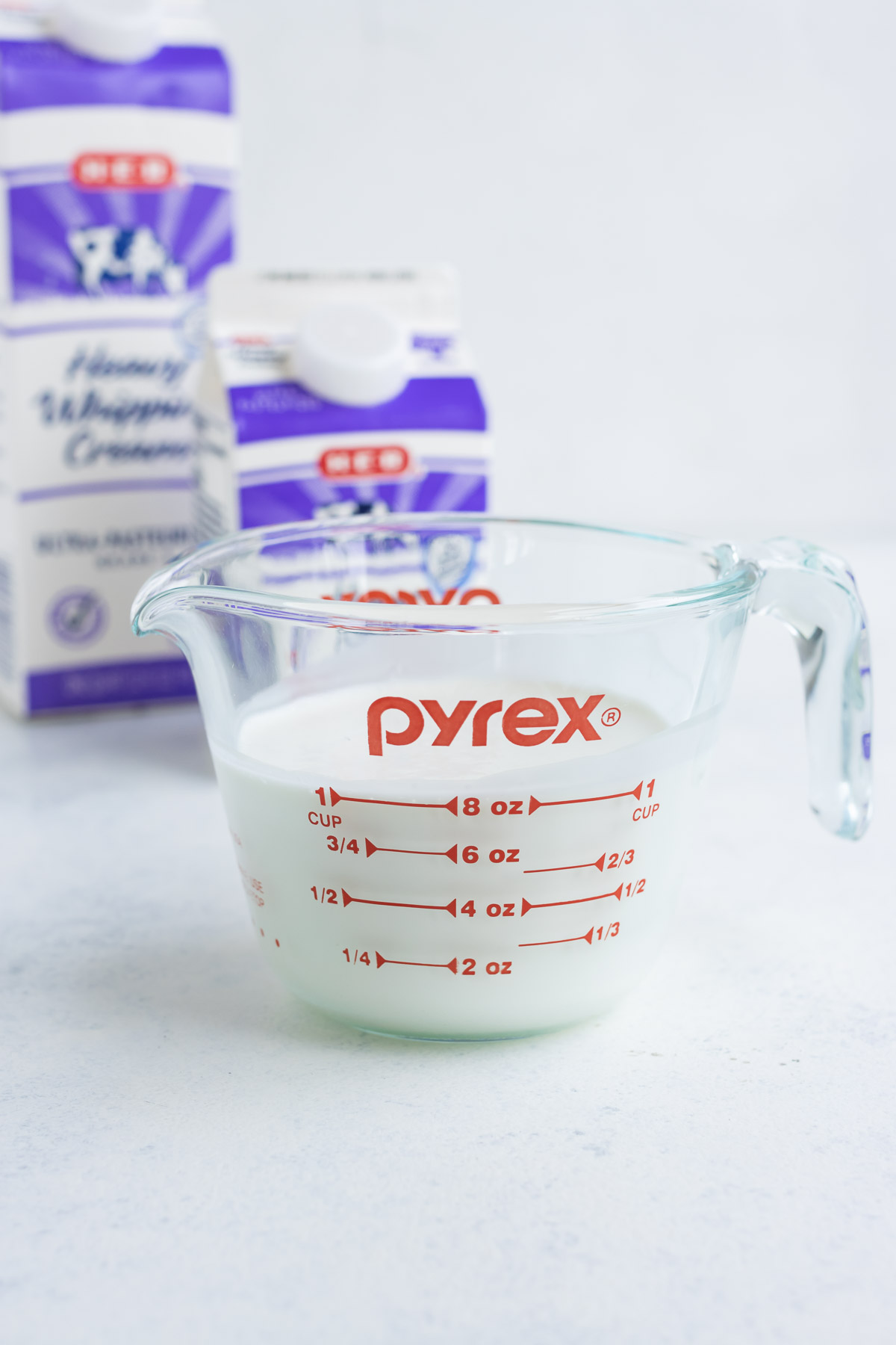 Heavy whipping cream poured in a glass measuring cup with the original containers in the background.