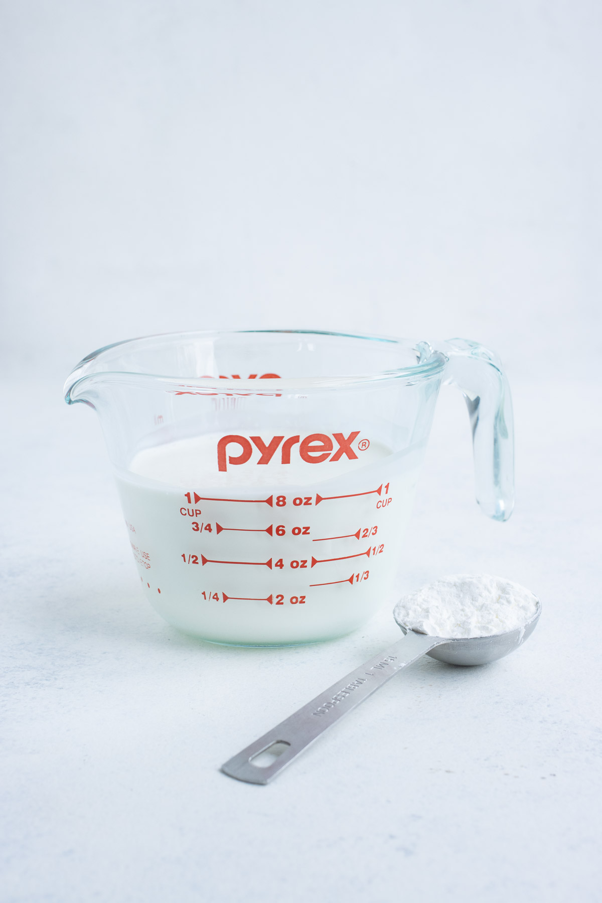 Milk in a glass measuring cup with a teaspoon of cornstarch on the side.