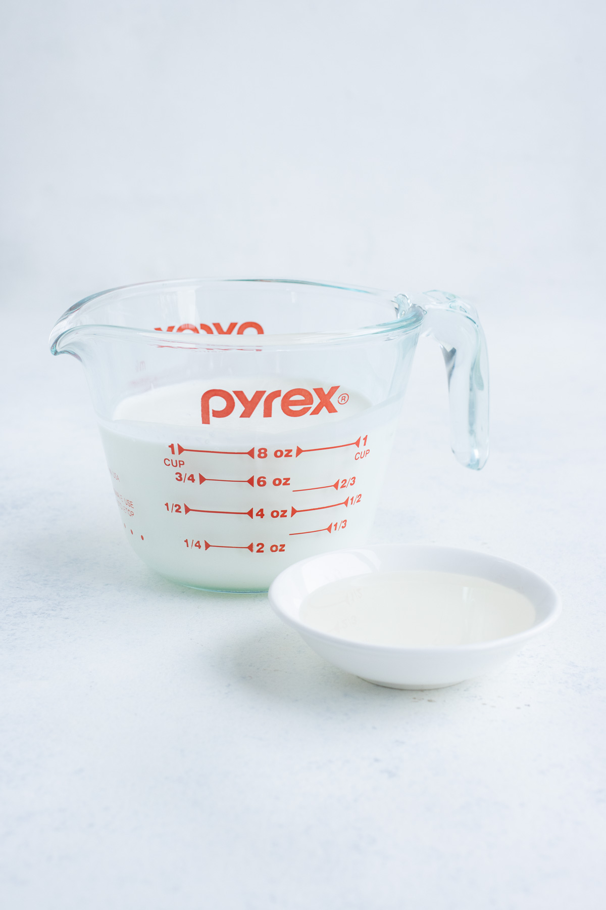 Milk in a glass measuring cup with a white bowl of vinegar on the side.