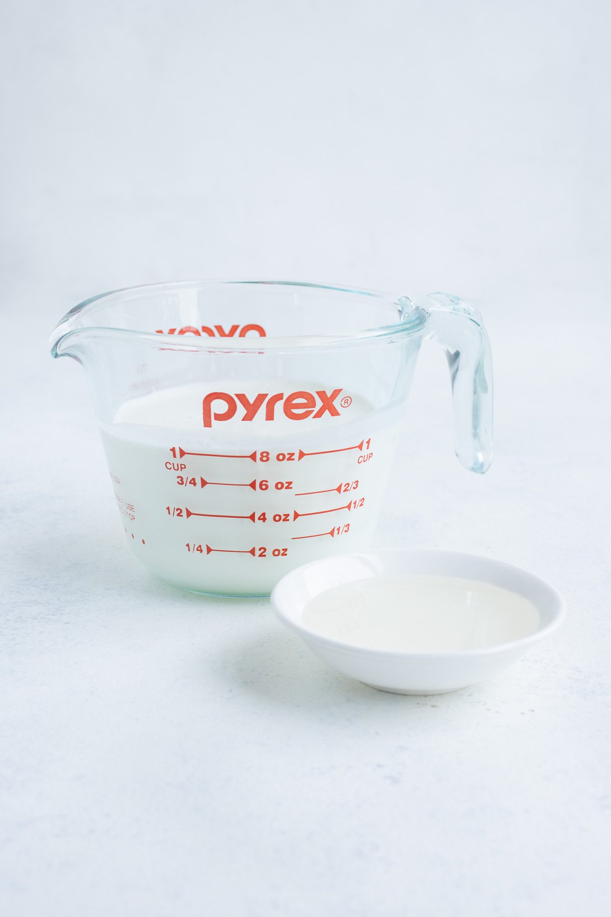 Milk in a glass measuring cup with a white bowl of vinegar on the side.