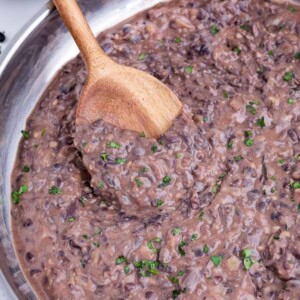 A wooden spoon stirs refried black beans in a skillet.