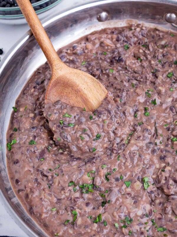 A wooden spoon stirs refried black beans in a skillet.