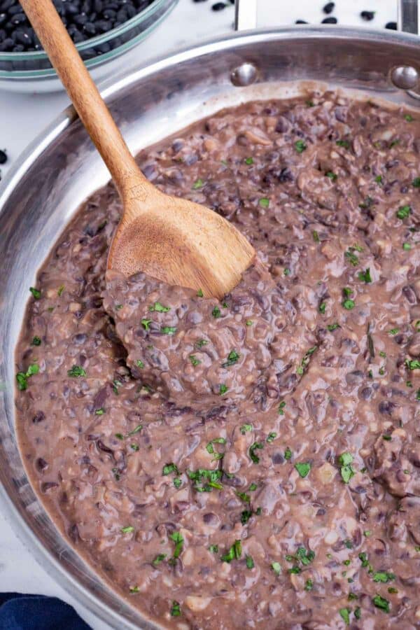 A wooden spoon stirs refried black beans in a skillet.