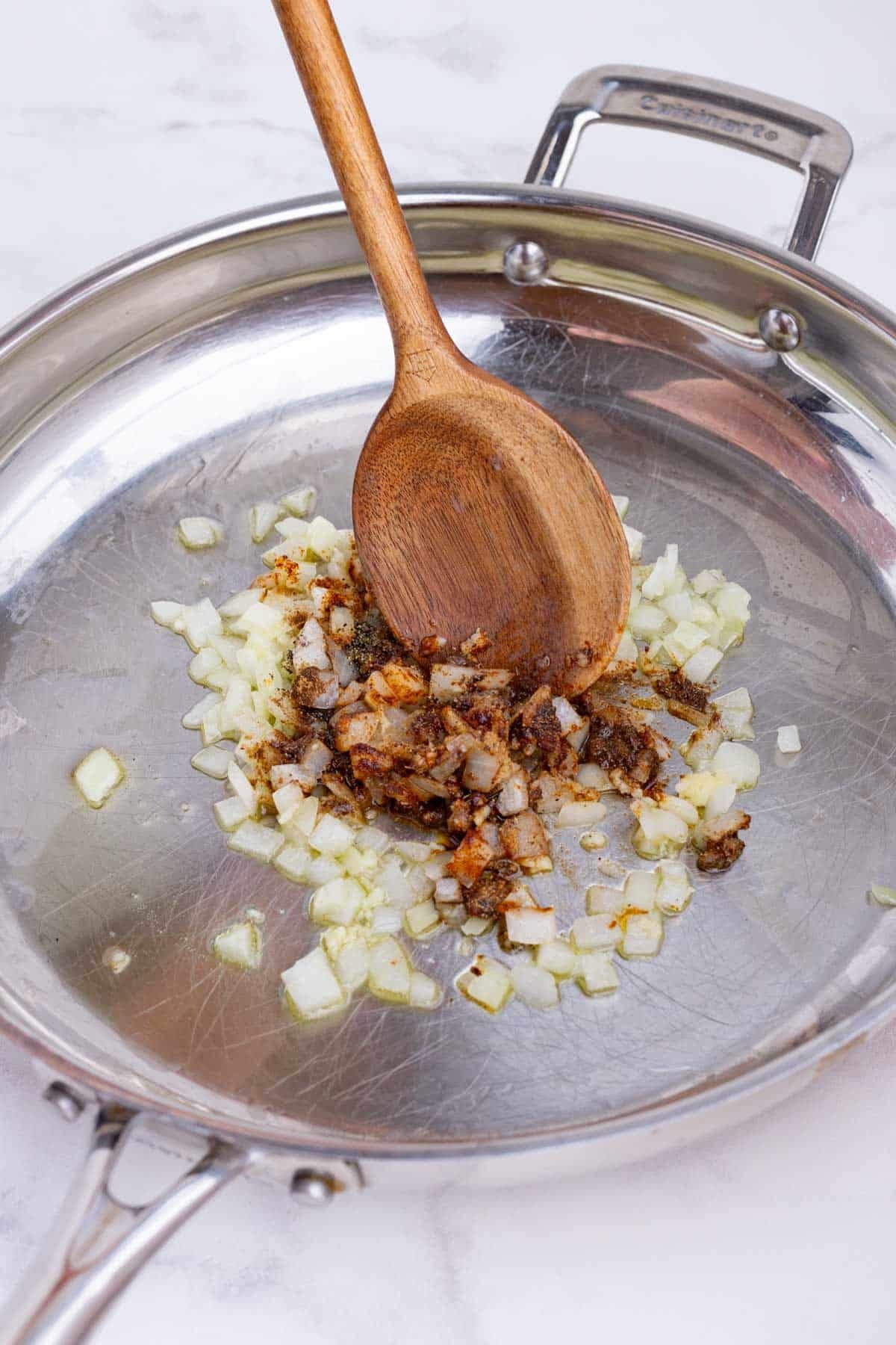 A spoon stirs the onion and seasonings in the skillet.